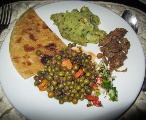 Plate with a folded flatbread, a scoop of mashed potatoes with herbs, a serving of cooked peas with diced vegetables, a small portion of browned meat pieces, and a garnish of chopped fresh herbs.