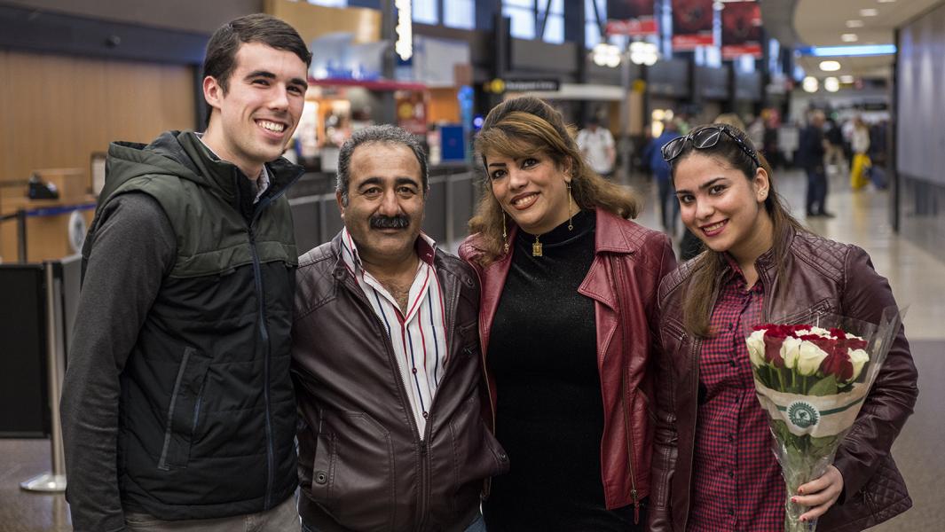 Four people stand closely together in an airport terminal, posing for a photo. The group is wearing jackets, and one person on the right is holding a bouquet of red and white roses wrapped in paper. The background shows airport signage, seating, and travelers walking through the terminal.