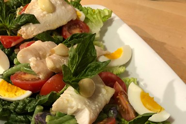A composed salad with pieces of cooked fish, tomatoes, green beans, lettuce, basil, and sliced hard‑boiled eggs on a white plate.