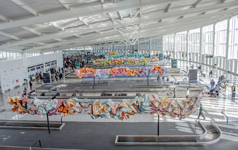 Large, bright airport baggage claim hall with colorful suspended artwork.