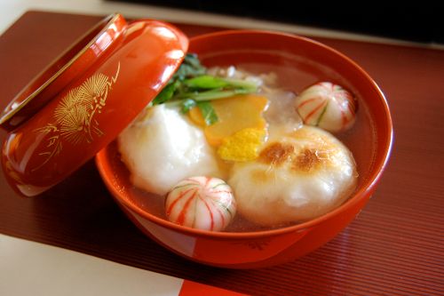 Red lacquer bowl of soup with toasted mochi pieces, leafy greens, and decorative red-and-white fish cake balls, with the lid resting partly open.