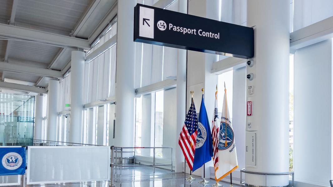 An airport interior with a sign pointing to passport control and several official flags displayed beneath it.