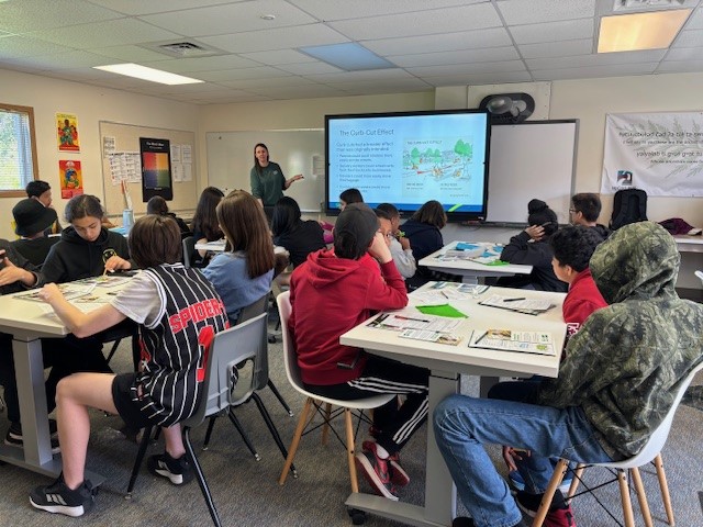 A classroom where students sit at tables looking toward a large screen as an instructor presents information.