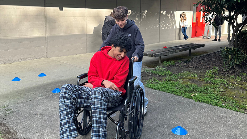 A person sits in a wheelchair while another person pushes it along an outdoor path marked with small cones.
