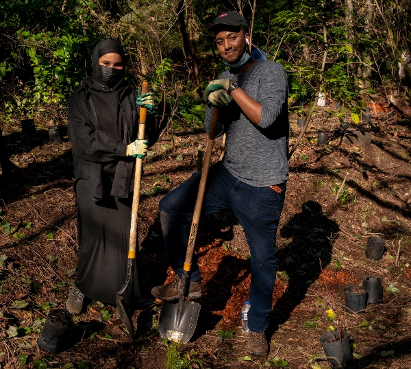 Two people standing in a wooded area holding shovels during an outdoor planting activity.