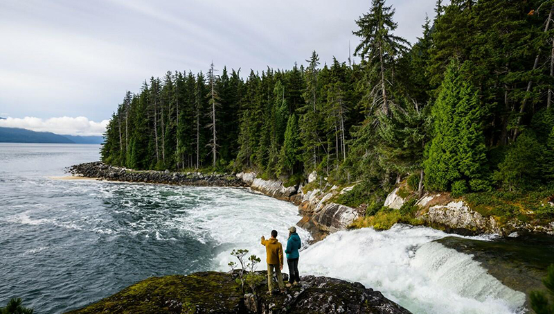 Two people stand on a rocky overlook beside a rushing waterfall leading into a coastal inlet surrounded by dense evergreen forest.
