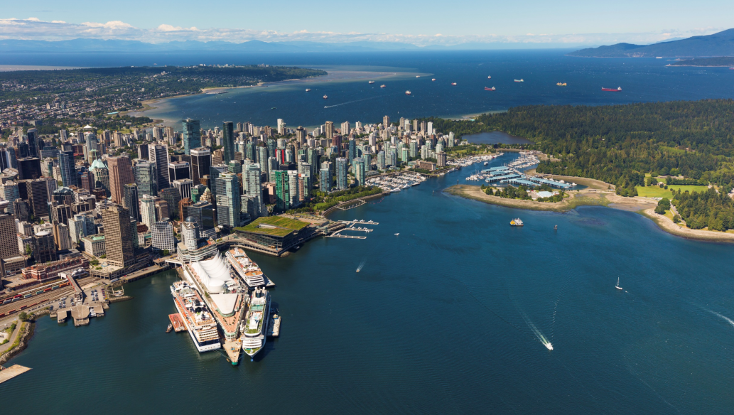 Aerial view of a coastal city skyline with cruise ships docked at a waterfront terminal and forested parkland along the shoreline.