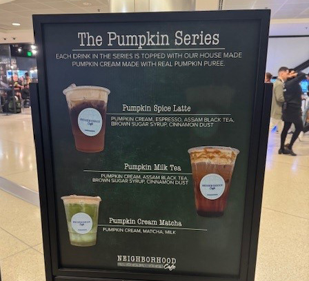 A standing sign titled “The Pumpkin Series” displaying three drinks labeled Pumpkin Spice Latte, Pumpkin Milk Tea, and Pumpkin Cream Matcha, each shown with the drink name and ingredients, located inside an airport café area.