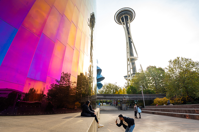 The colorful reflective exterior of MoPOP with the Space Needle rising in the background and several people in the surrounding plaza.