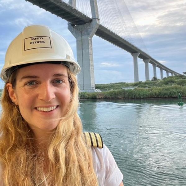 Rachel Pagonis wearing a hard hat labeled 'Safety Officer' stands near the water with a large bridge in the background.