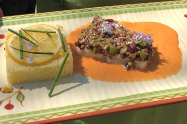 A plated dish with a crusted fish filet on an orange sauce and a square of cornbread topped with lemon slices and chives.