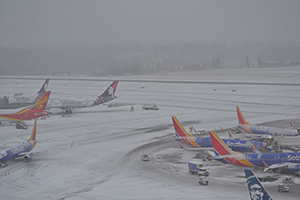 A snowy airport scene showing multiple airplanes parked at gates during low-visibility winter weather.