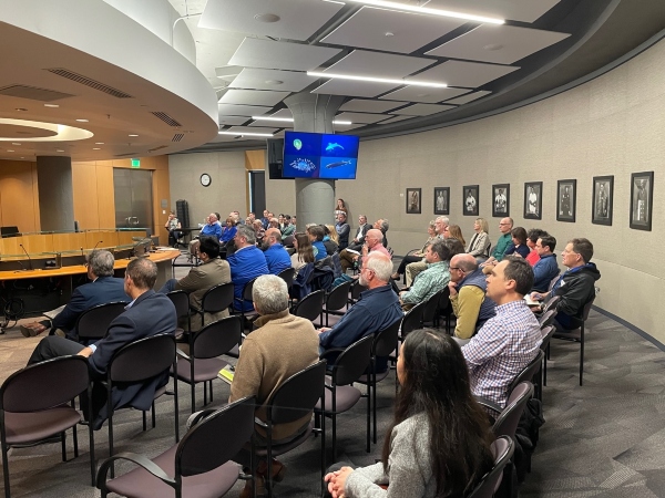 A group of people sit in rows of chairs inside a modern meeting room, facing a presentation area with two screens displaying images of fish. The room has curved architectural features, framed photographs on the wall, and a presenter standing near the front addressing the audience.