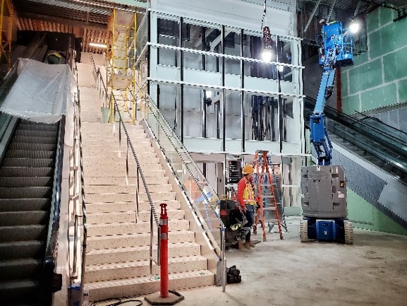 Construction area with a central staircase, two escalators, scaffolding, and workers installing equipment using a lift.