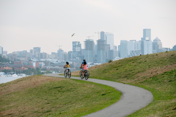 Two people ride bicycles along a winding path on a grassy hill with a city skyline in the background.