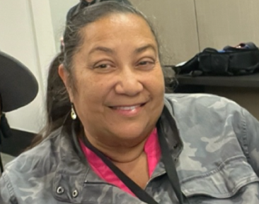 Headshot of Shannon Acena-Neal seated indoors, wearing a patterned jacket, with a table and bags in the background.