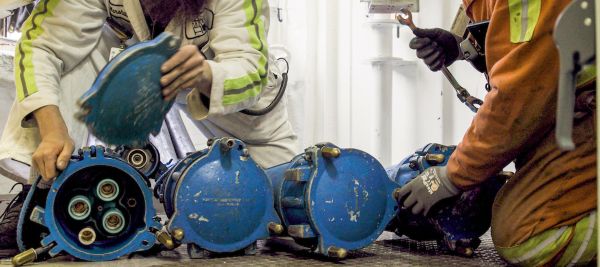 Two workers handle and inspect several large blue industrial connectors on the floor inside a work area.