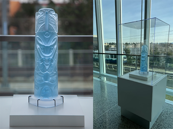 A tall, light‑blue carved glass totem‑style sculpture displayed on a white pedestal, shown both in a close view and inside a clear protective case near large windows.