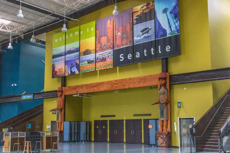 A large “Welcome to Seattle” display hangs above the entrance area inside a conference or terminal building, with colorful panels and carved wooden figures on each side.