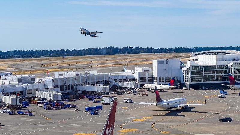 An airport terminal with multiple airplanes on the tarmac, ground service vehicles, and a plane taking off in the background.