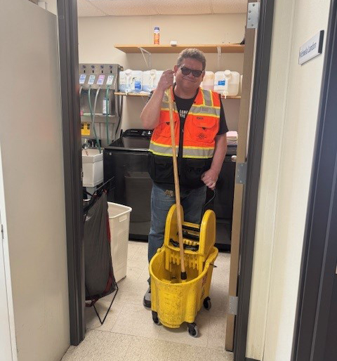Todd Nuttman wearing a high‑visibility safety vest stands in a custodial room holding a mop in a yellow mop bucket.