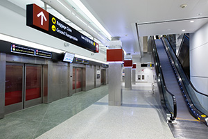 Modern airport corridor with an escalator and automated transit doors, with overhead signs for baggage claim and ground transportation.
