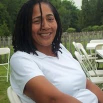 Virginia Fullwood outdoors on a folding chair, wearing a light-colored shirt, with other chairs and a wooden fence in the background.