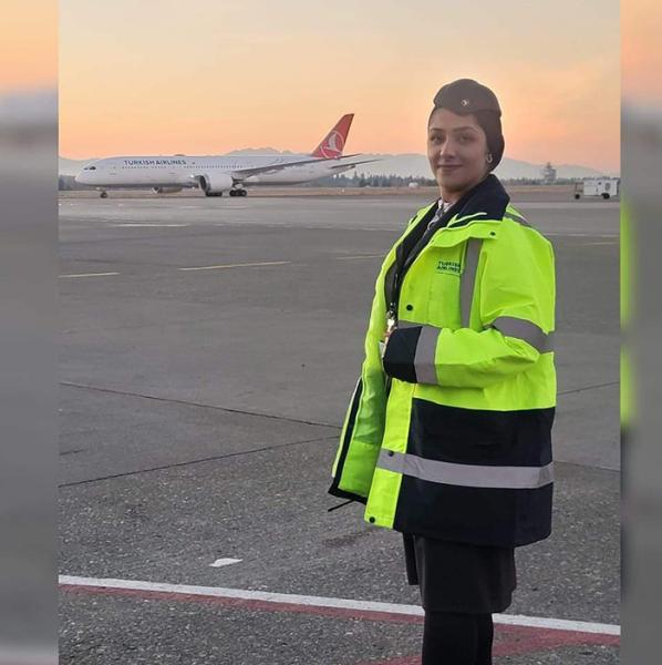 Zainab Hasan in a high‑visibility jacket stands on an airport tarmac with an airplane in the background at sunset.