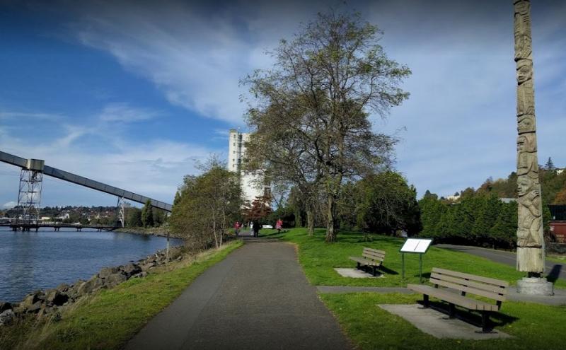 A paved waterfront path runs alongside a rocky shoreline and grassy park area with benches, trees, and a tall carved wooden totem pole. A few people walk in the distance, and an elevated industrial conveyor structure extends over the water under a blue sky with scattered clouds.