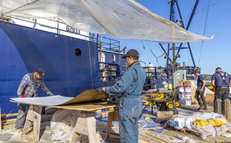 Several people working outdoors on a ship‑side project area, handling large materials and equipment under a temporary plastic canopy.