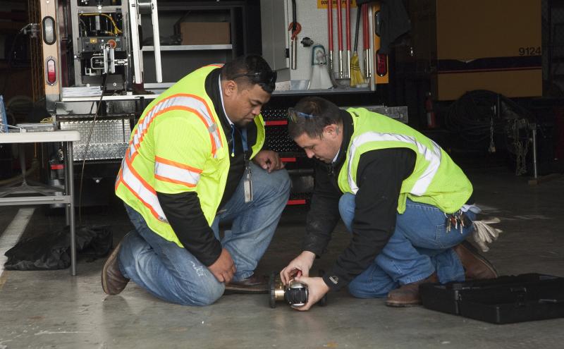 Two workers wearing safety vests and protective gear are kneeling on a concrete floor beside a utility vehicle, examining a small wheeled inspection device placed between them. Tools, equipment, and storage compartments are visible in the background.