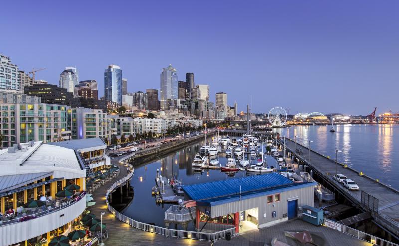 Evening view of a waterfront marina with several docked boats, a curved boardwalk with outdoor dining areas, and a city skyline of tall buildings in the background. A large Ferris wheel and illuminated structures sit along the shoreline, with calm water reflecting lights from the surrounding area.