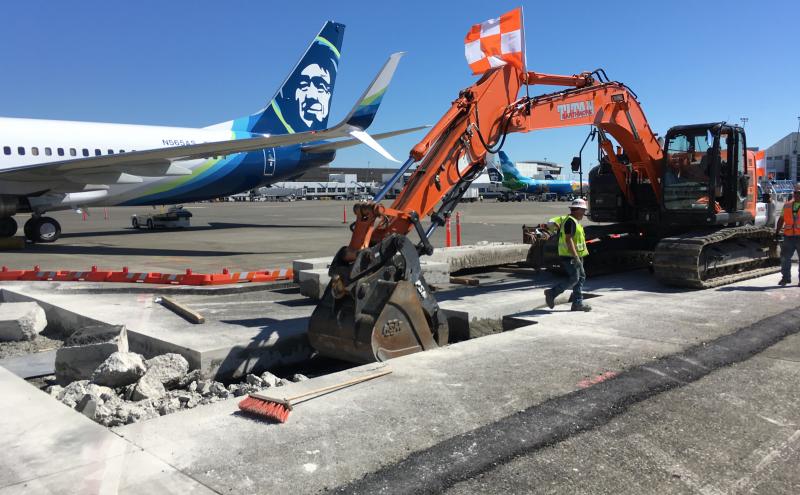 Construction equipment and workers breaking up concrete on an airport tarmac near a parked airplane, with debris and tools visible around the work area.