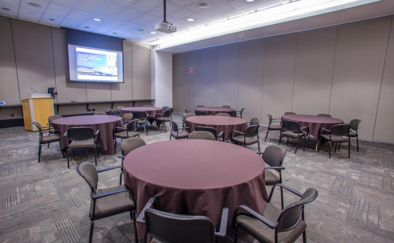 View from the entry doorway looking into the Beijing Conference room. The room is set with Round tables with chairs, and each table is covered in a brown tablecloth. The projector is turned and the projector screen is displaying content