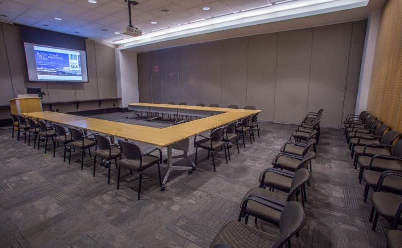 View from the entry doorway looking into the Beijing Conference room that is in a u-shape room set and two rows of theater style seating behind it. The projector is turned and the projector screen is displaying content