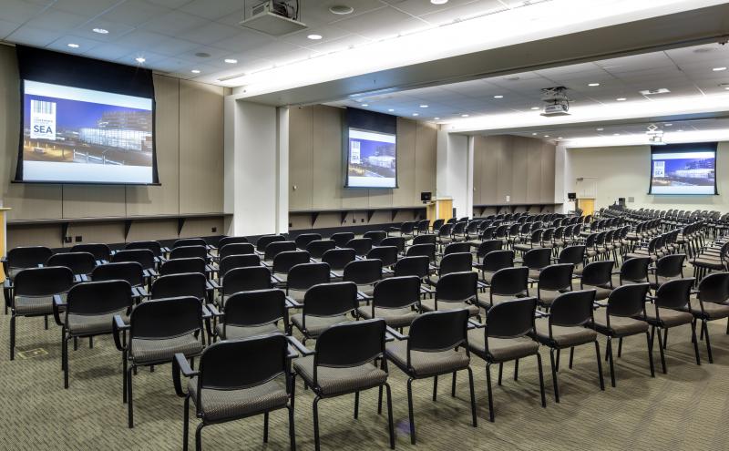 View of the International Auditorium conference room from the left Beijing entry doorway. The room is set in theater style seating for 210 people. All Airwalls are open, all three projectors are turned on, and all three dropdown projectors screens are displaying content.