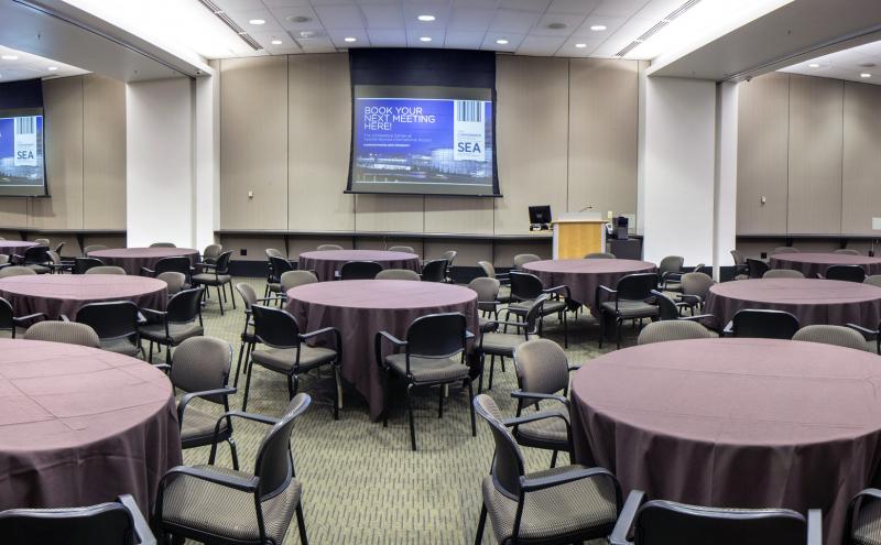 Panoramic view of the International Auditorium conference room. The room is set in 25 banquet rounds, with 7 chairs each. There are brown table cloths on each round table. All Airwalls are open, all three projectors are turned on, and all three dropdown projectors screens are displaying content.