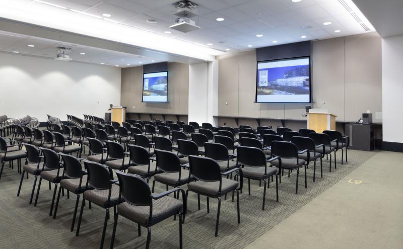 View from the London entry doorway looking into the International B conference room. The room is set theater style for 144 people facing the front of the room. Two projectors are turned and both projector screens are displaying content.