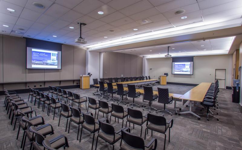 View from the London entry doorway looking into the International A conference room. The room is set in a u-shape, with rows of theater style seating behind it, facing the Amsterdam dropdown screen. Two projectors are turned and both projector screens are displaying content