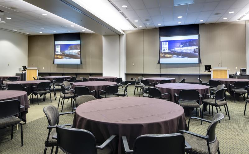 View from the London entry doorway looking into the International B conference room. The room is set with Round tables with chairs, and each table is covered in a brown tablecloth. Two projectors are turned and both projector screens are displaying content.