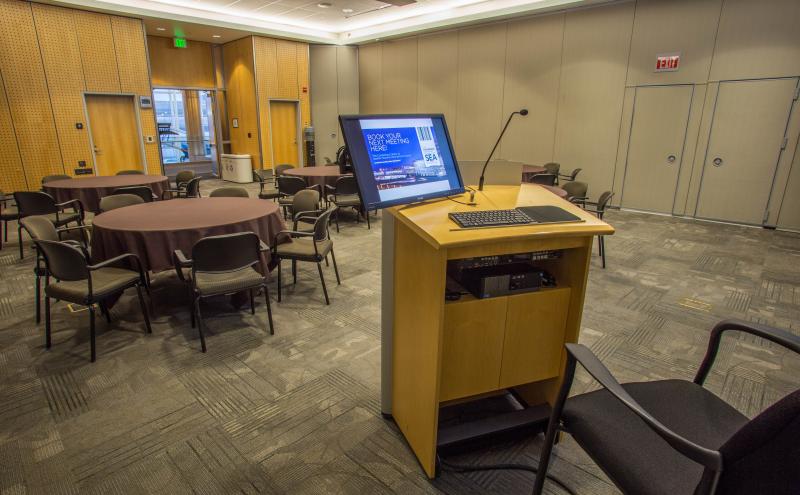 View from the podium located inside the London Conference room featuring 6 banquet round tables with brown table clothes and chairs.