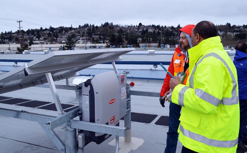 Two workers in safety gear examine rooftop solar equipment at Fishermen’s Terminal, with solar panels and surrounding buildings visible under an overcast sky.