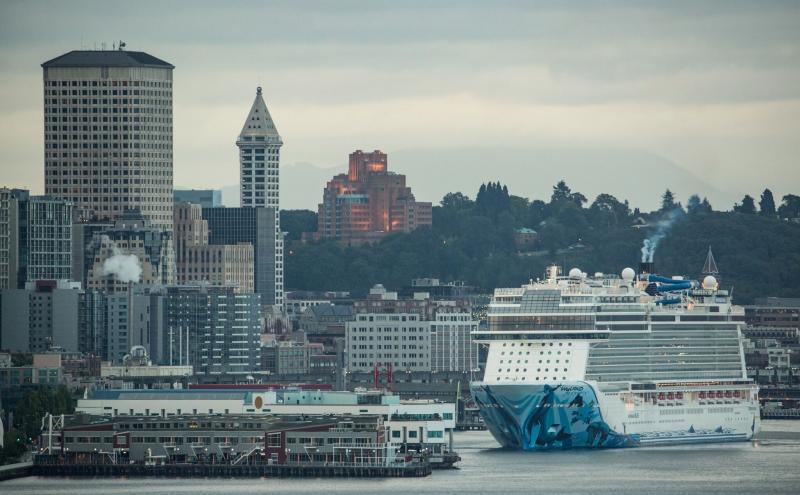 A large cruise ship approaches the Seattle waterfront, with downtown buildings and a cloudy sky in the background.