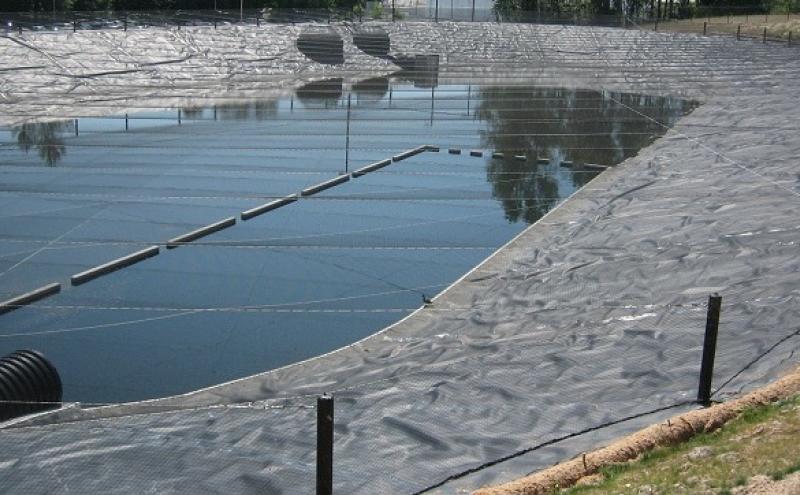 Large outdoor containment pond covered with a protective mesh or liner, with support poles and tensioned cables running across the surface. Surrounding the pond is a sloped embankment with vegetation and a fence.
