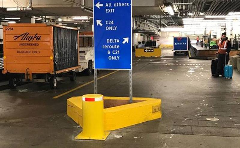 Underground airport service area with a large blue directional sign showing exits and baggage cart traffic routes, surrounded by baggage carts, equipment, and a worker standing near luggage.