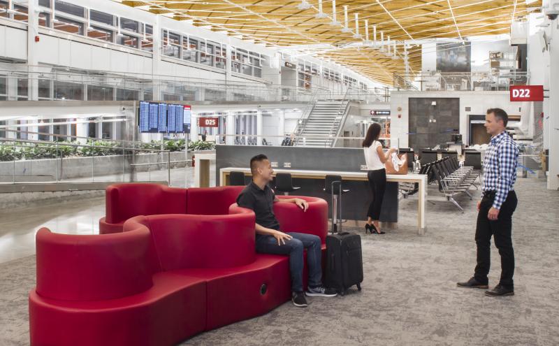 Interior of Concourse D Annex at SEA showing curved red seating, a gate agent podium and travelers near gates D22 and D25. 
