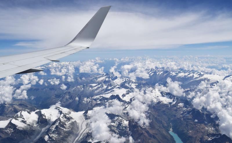 View from an airplane window showing the wing above snowy mountain peaks and scattered clouds.