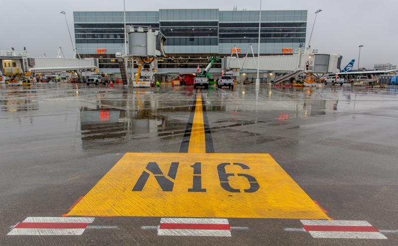 A wet airport tarmac showing a large yellow N16 marking leading toward terminal gates with jet bridges and ground vehicles.