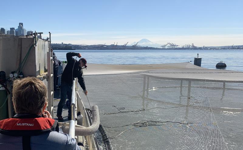 A person standing on a shoreline near metal equipment while another person on a boat looks toward the water on a clear day.