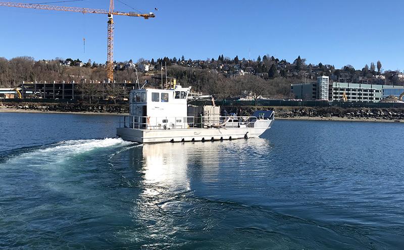 A small workboat moves across calm water near a shoreline with buildings, trees, and a construction crane in the background on a clear day.
